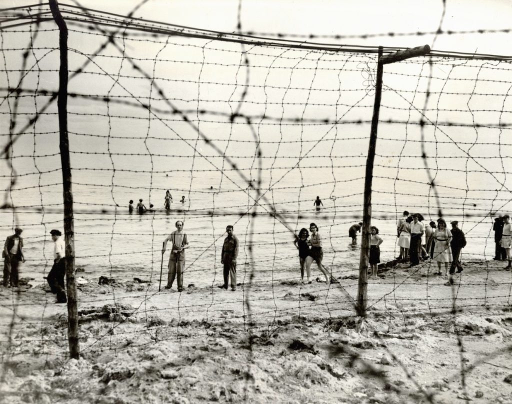 A group of Jewish refugees in a British detention camp in Cyprus swim in the Mediterranean Sea behind a barbed wire fence that surrounds the camp, 1946. The refugees were permitted use of the beach from 10 a.m. to 5 p.m. daily. (Courtesy JDC Archives)