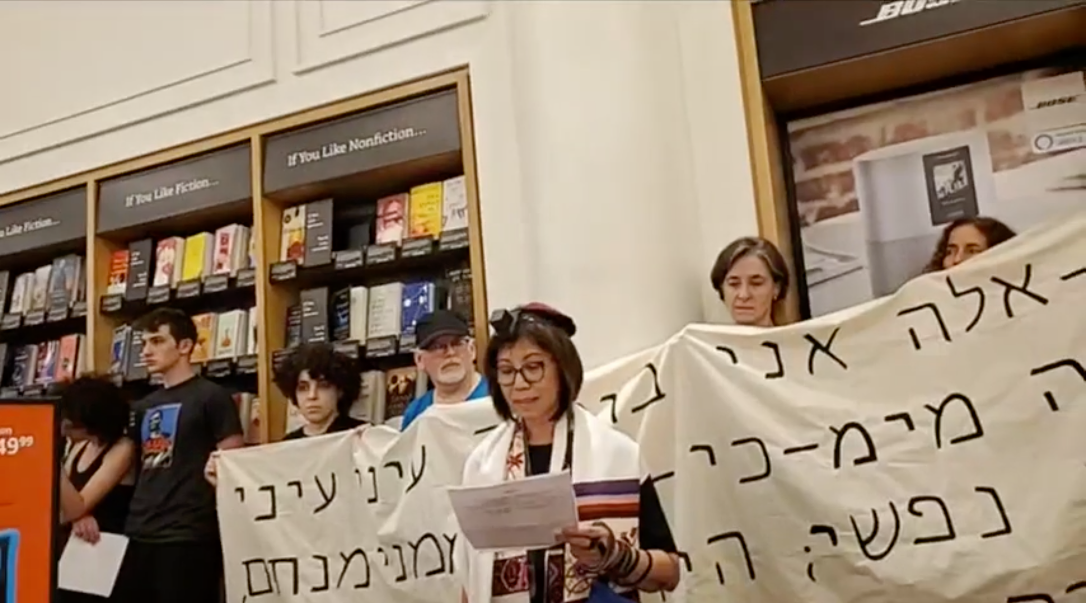 A demonstrator speaks at a Jewish protest against Amazon at one of the company's brick-and-mortar stores in New York City on Sunday, August 11. The protest was held on Tisha B'Av, a traditional Jewish day of mourning. Behind the speaker is a banner with a Hebrew quotation from the Book of Lamentations. (Screenshot from Facebook)