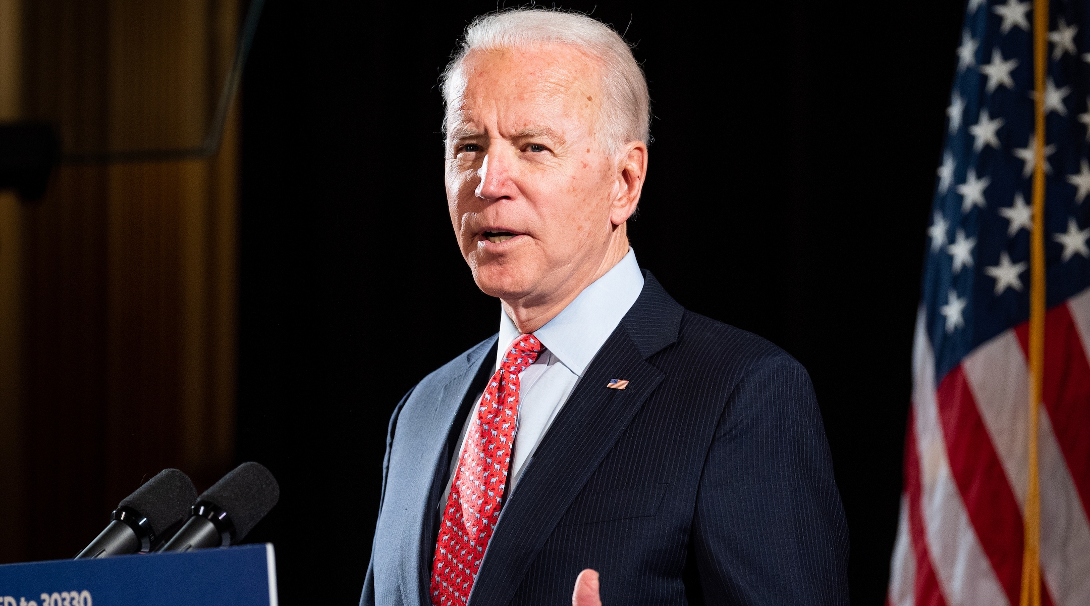 Former vice president Joe Biden-- a white man in his late 70s with silvery hair wearing a navy suit and red tie-- speaker before a podium with an American flag in the background