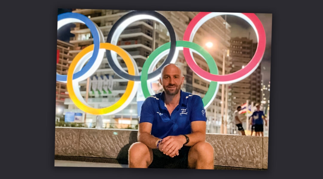 A man sitting at the Olympic Games in Tokyo, the Olympic rings behind him.