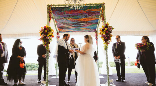 A Jewish couple marries outdoors underneath a colorful chuppah