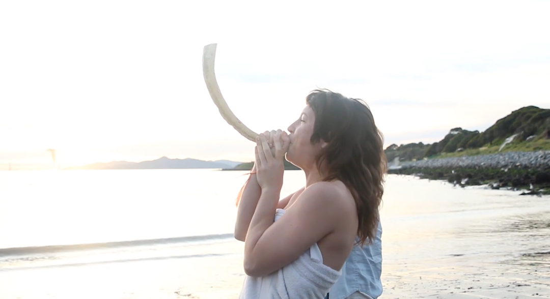 A woman blowing a shofar along a bay