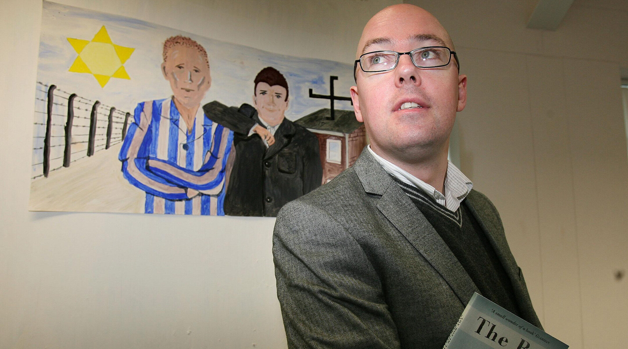 Man poses with book in front of children's drawing of Auschwitz