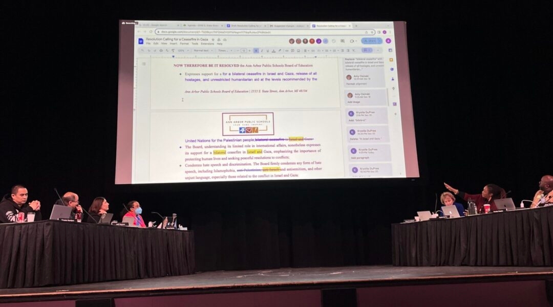 A view of a school board meeting on a stage with a giant Google Doc projection overhead