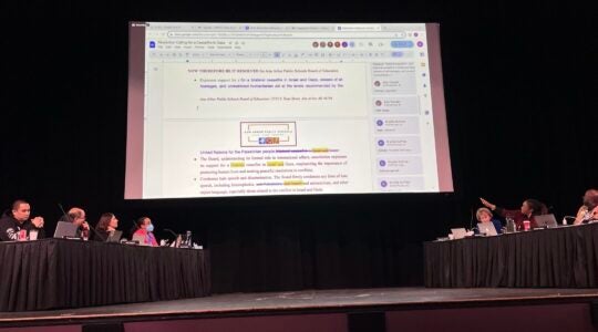 A view of a school board meeting on a stage with a giant Google Doc projection overhead