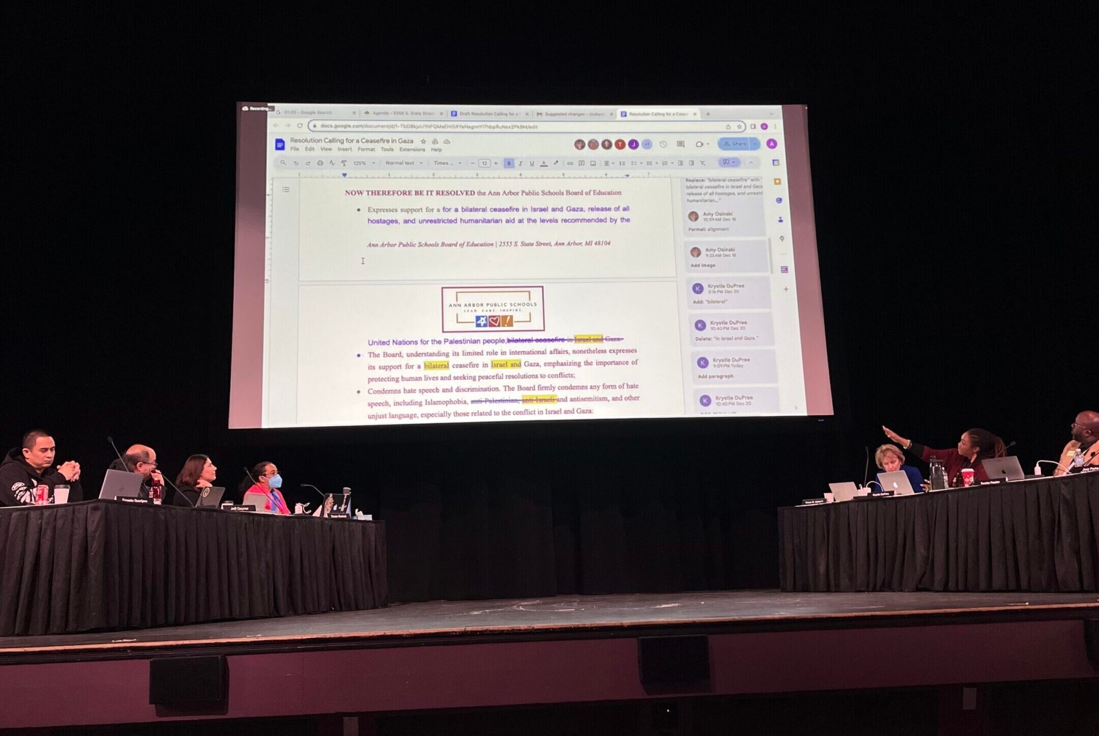 A view of a school board meeting on a stage with a giant Google Doc projection overhead