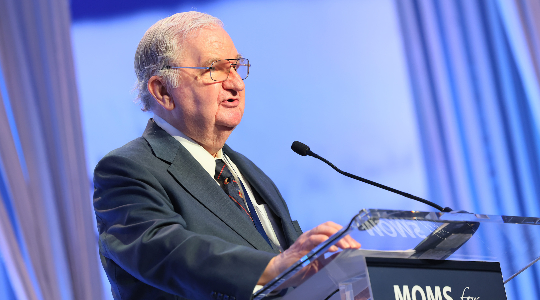 A man delivers a lecture at a podium labeled "Moms For Liberty"