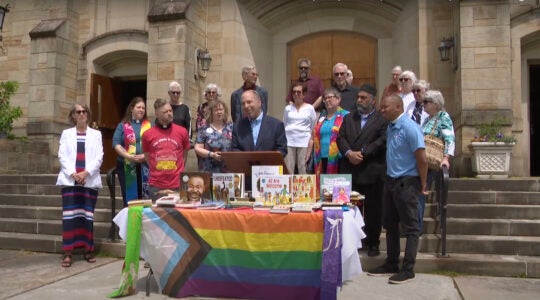 A rabbi speaks at a press conference in front of a table stacked with books and the LGBTQ flag