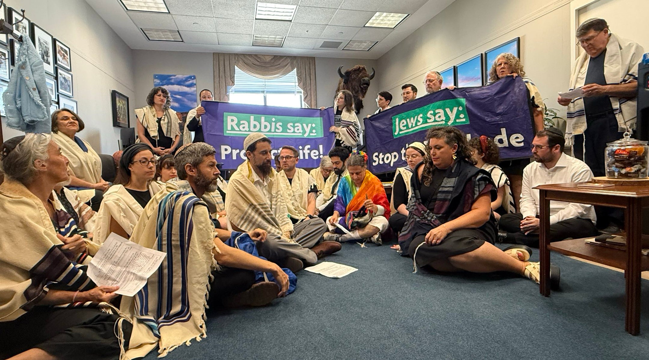 A group of rabbis sit and stand in Sen. John Thunes office, holding banners that read "Rabbis say: Protect Life!” and “Rabbis say: Stop the Blockade."