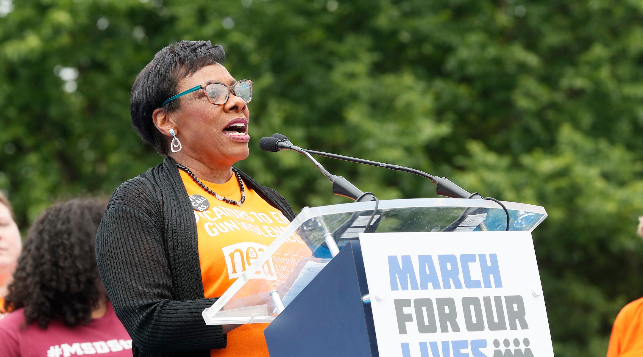 NEA president rebecca pringle speaking in an orange shirt at a podium.