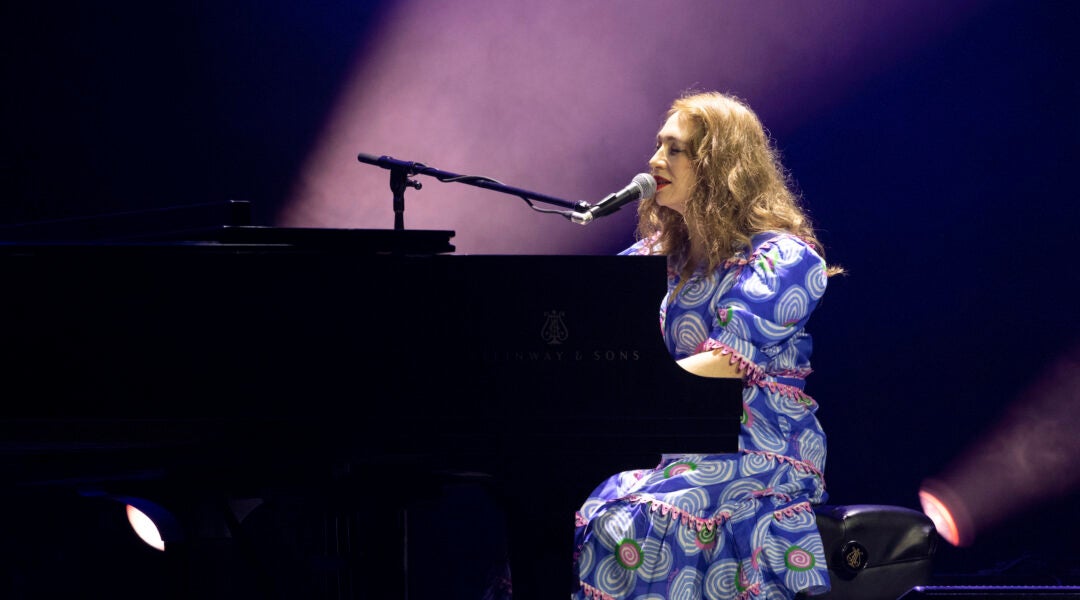 A photo of regina spektor in a purple dress playing a piano and singing into a mic.