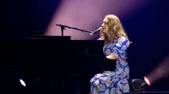 A photo of regina spektor in a purple dress playing a piano and singing into a mic.