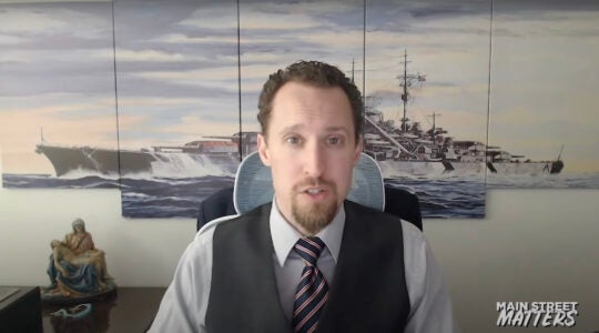 A man in a vest and tie interviewed in front of art depicting a Nazi warship