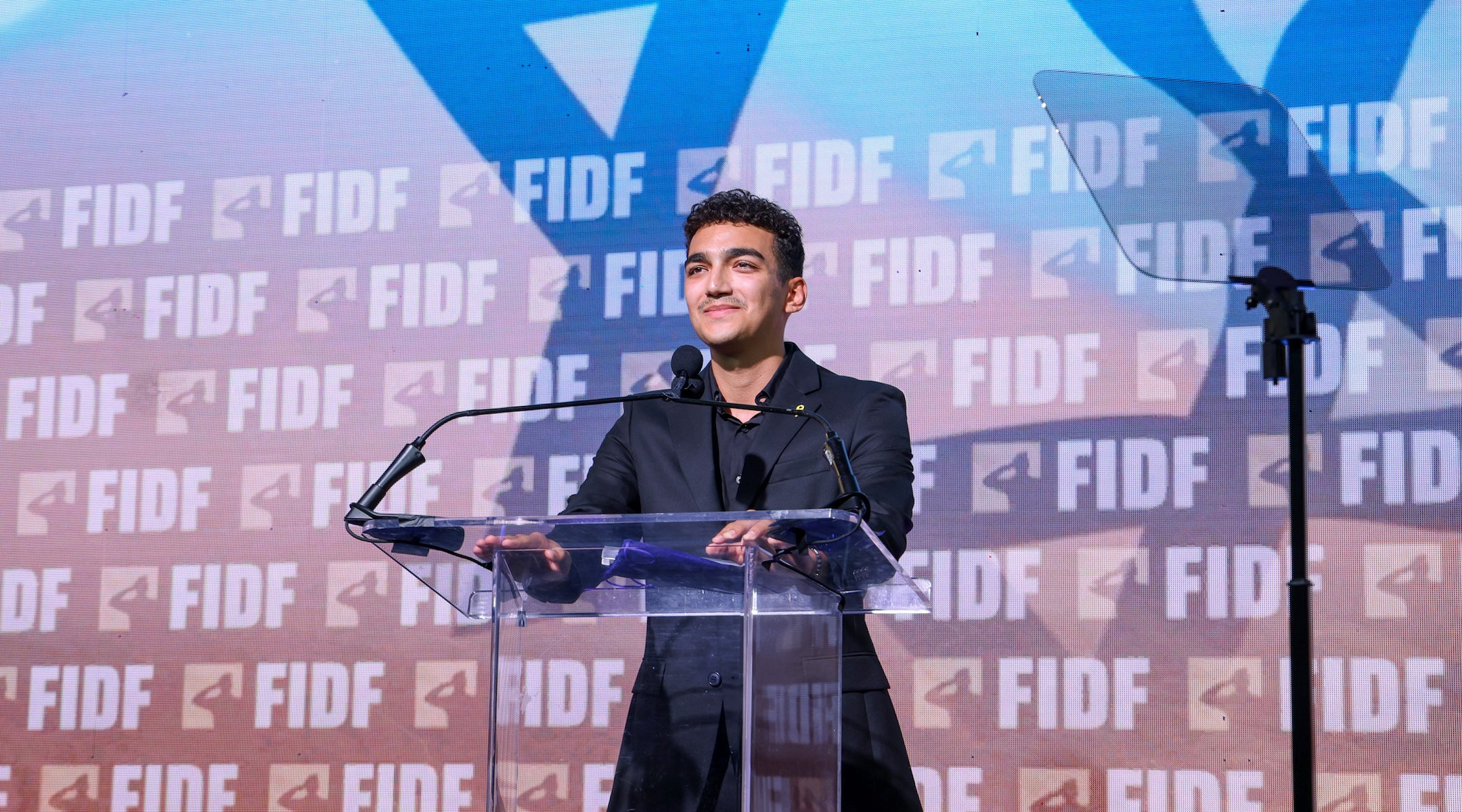 A photo of Edan Alexander speaking from a podium.