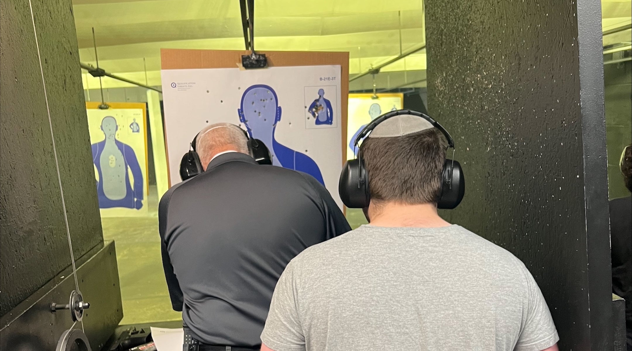 two people facing away from the camera look at a target at a gun range.