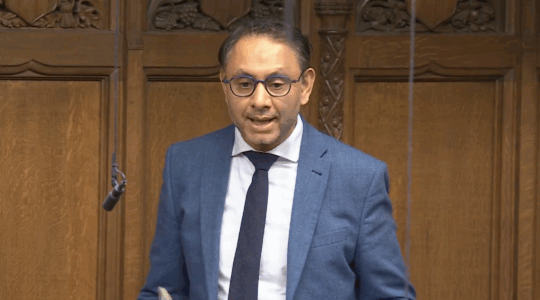 A photo of Shockat Adam speaking at parliament in a blue suit.