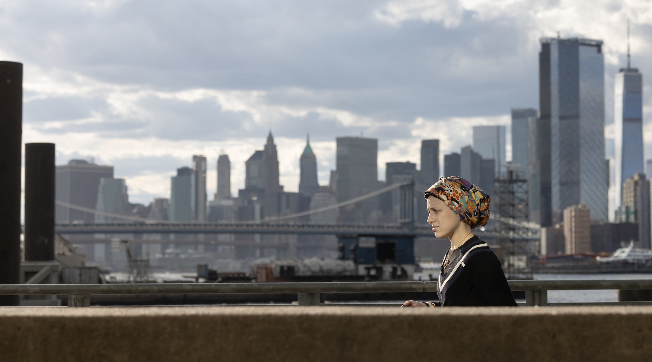 A photo of a Hasidic woman walking across a bridge with Manhattan in the background.